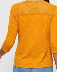 Vrouw in een mosterdgele 3/4 mouw top met sheer stippenstof over de bovenrug, gecombineerd met lichtblauwe jeans.