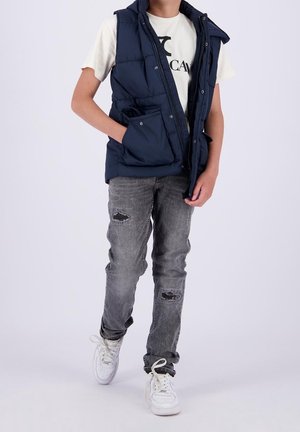 Marineblauwe puffer vest met rits en zakken, gedragen over een wit T-shirt, gecombineerd met versleten grijze jeans en witte sneakers.