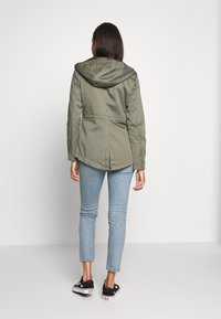 Kvinna som står med ryggen mot kameran, iklädd en olivgrön huvtröja, ljusblå jeans och svarta sneakers mot en enfärgad bakgrund.