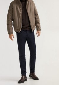Brown bomber jacket, dark sweater, navy pants, and brown ankle boots. Smooth materials with elastic cuffs; fitted design with minimal detailing.