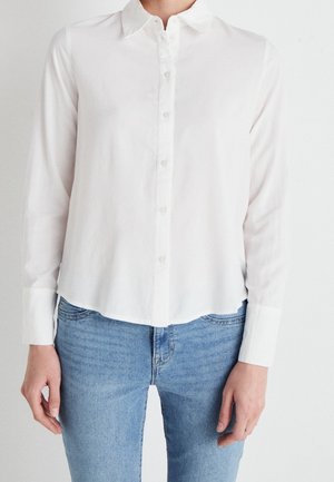 Personne portant une chemise blanche à manches longues boutonnée et un jean bleu clair, debout devant un fond blanc uni.