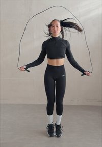 Top court noir ajusté à col haut, associé à un legging noir taille haute et des chaussettes blanches. Tenant une corde à sauter, portant des baskets de sport.