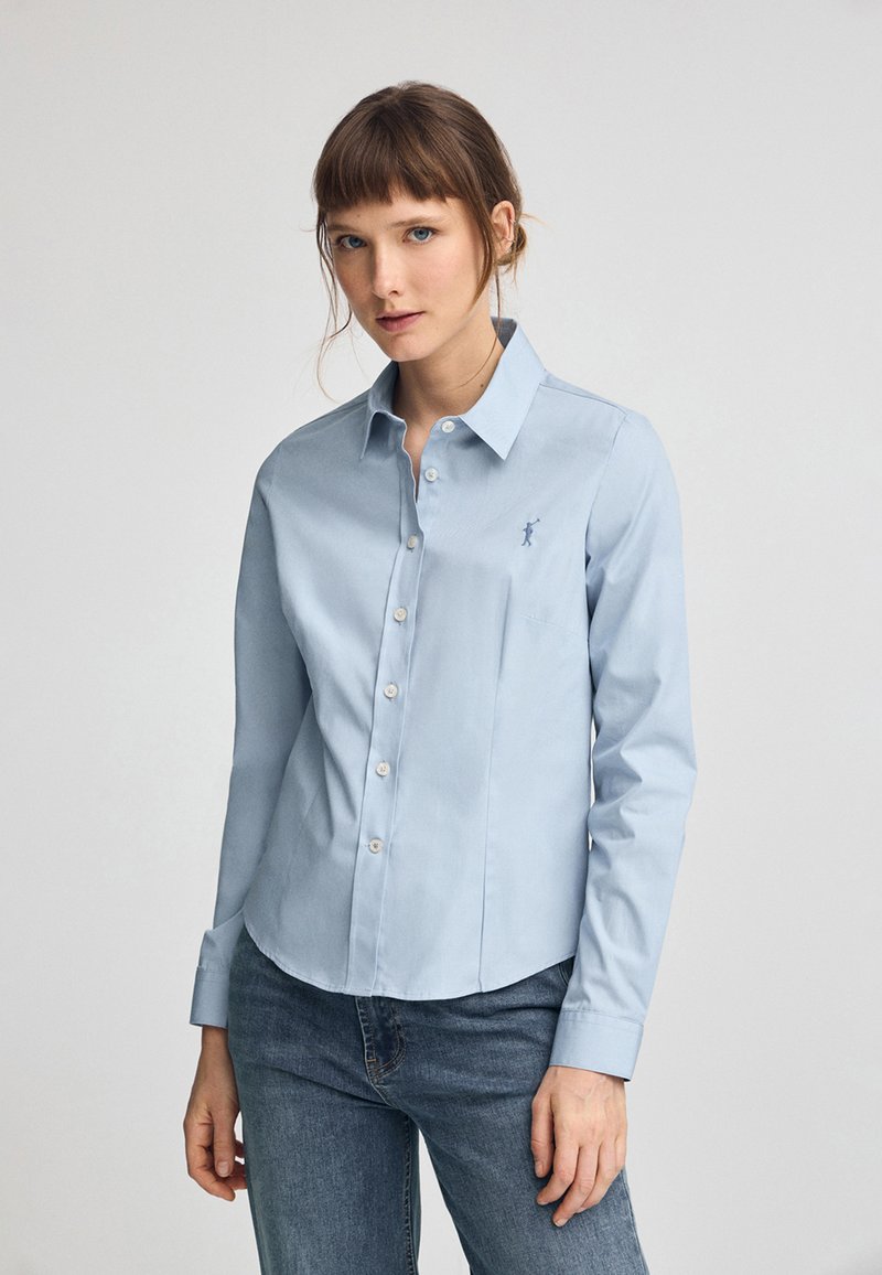 Camicia azzurra chiaro con bottoni, maniche lunghe, colletto a punta e un piccolo logo ricamato sul petto. Realizzata in un tessuto morbido e tessuto.
