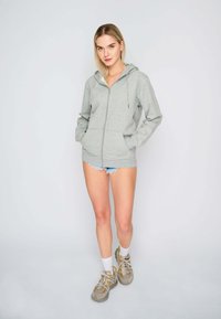 Felpa zip-up grigia chiara con tasca frontale, cappuccio con coulisse e texture morbida, abbinata a pantaloni shorts in denim e sneakers chunky.