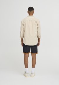 Beige langermet skjorte med rullede mansjetter, kombinert med marineblå shorts med brun kant. Brukt med hvite sokker og kremfargede sneakers.
