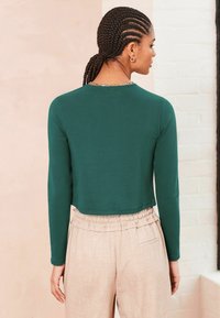Zelený dlhý rukáv cropped top s okrúhlym výstrihom, kombinovaný s béžovými vysokými, textúrovanými nohavicami s elastickým pásom.