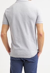 La chemise polo grise à manches courtes présente un col plat et un ourlet droit. Le tissu semble lisse avec une texture subtile et un design ajusté.