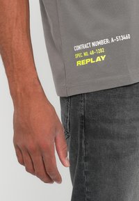Vue de profil d'une personne portant un t-shirt gris avec les textes "CONTRACT NUMBER: A-513460" et "REPLAY" en jaune, assorti à un jean gris foncé.