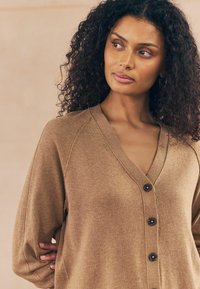 Cardigan en tricot marron avec un col en V, quatre boutons noirs et des manches raglan avec des détails de couture visibles, fabriqué à partir d'un tissu doux.