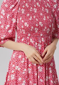 Robe florale rouge avec un corsage smocké et des manches bouffantes. Le tissu a un motif texturé avec de petites fleurs blanches et des accents bleus.