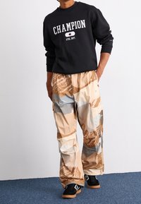 Black sweatshirt with "CHAMPION" logo, paired with beige and brown patterned cargo pants. The pants have a loose fit and visible pockets. Black sneakers.