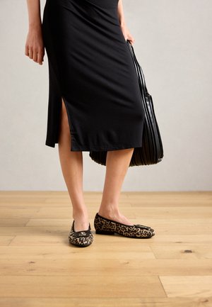 Nais, kes kannab musta midi-seelikut külgsuuna lõhega, hoides musta pleeditud kotti ja leopardimustriga lamedaid kingi puidust põrandal.