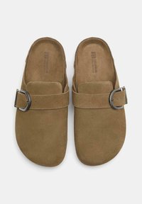 Tan ruskind slip-on sko med en rundt tå, prydet med en strop og metalspænde. Tekstureret indersål for komfort.