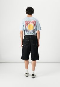 Una camiseta gráfica de color azul claro presenta un diseño de sol y montaña. Combinada con pantalones cortos de mezclilla oscura y calcetines blancos, muestra un atuendo casual.