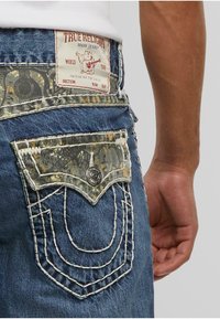 Tamno plave traperice s prepoznatljivom oznakom True Religion, s teksturiranom kamuflažnom stražnjom džepom, bijelim šavom i metalnim zakovicama.