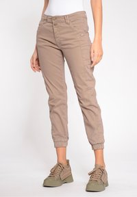 Pantalones cáqui tapered con una textura suave, puños elásticos, bolsillos frontales y costuras visibles. Combinados con zapatillas verde olivo.