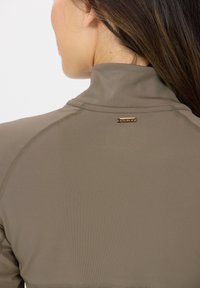 Taupe Sporttop mit hohem Kragen, aus glattem Stoff und einem kleinen goldfarbenen Logo nahe dem Hals. Nahtdetails sind sichtbar.