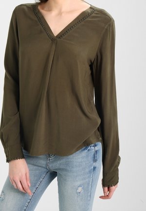 Blouse à manches longues couleur vert olive avec un col en V et des coutures décoratives le long de l'encolure et des poignets, fabriquée en tissu léger.