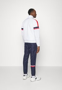 Hvitt jakke med glidelås og røde og marineblå striper, sammen med marineblå joggebukser. Har elastiske mansjetter og ankelsømmer, brukt med hvite sneakers.
