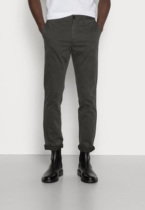 Hombre con pantalones gris oscuro remangados y botas negras de cuero hasta el tobillo, de pie con las manos en los bolsillos sobre un fondo blanco liso.