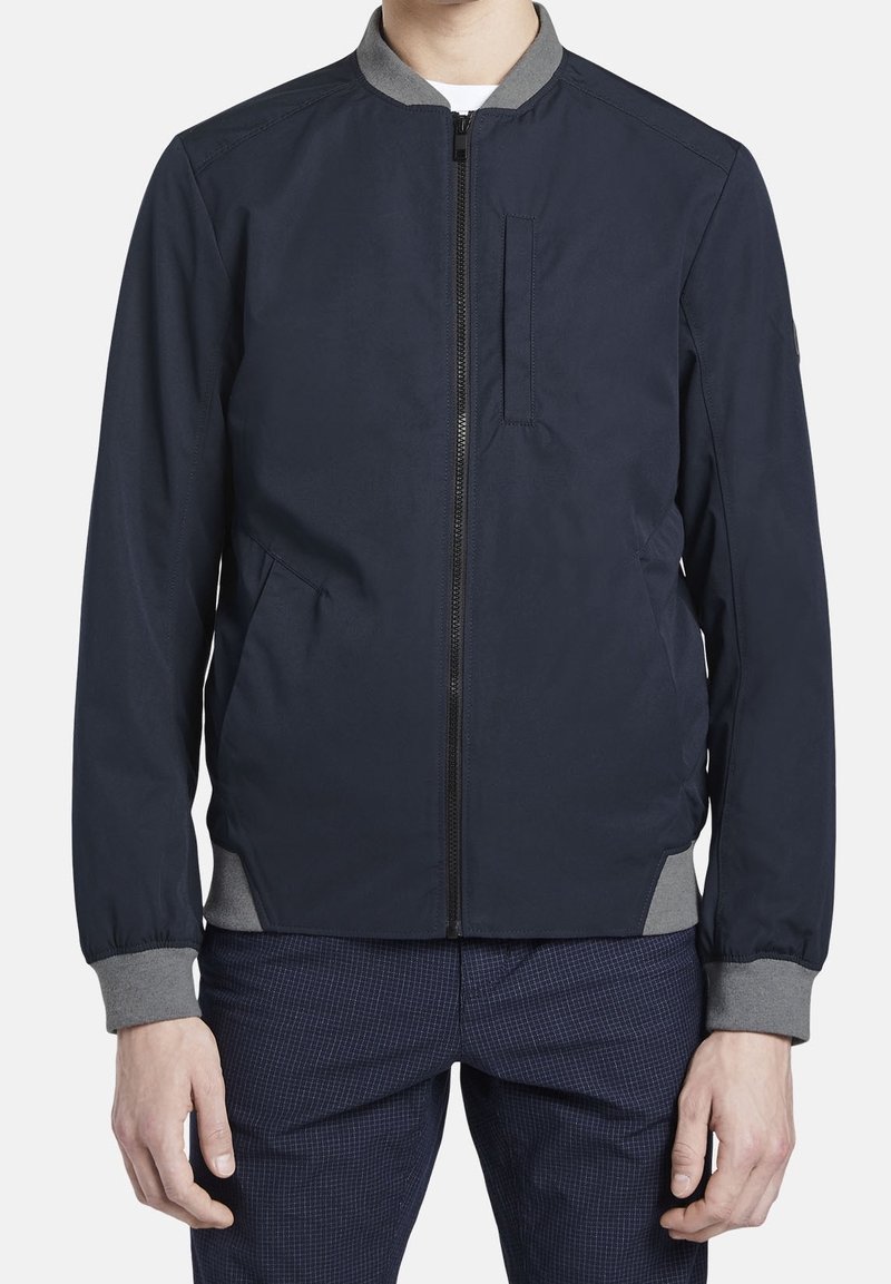 Námořnická bombarďák s šedým límcem, zipem v přední části, bočními kapsami a žebrovanými manžetami. Hladká tkanina s jednoduchým, moderním designem.