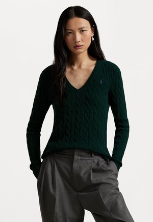 Pull en tricot vert foncé à col en V profond et à manches longues, arborant un petit logo sur la poitrine. Associé à un pantalon gris taille haute.
