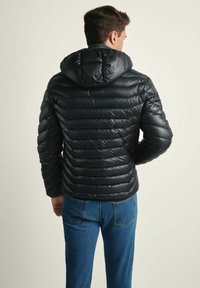 Czarna, puszysta kurtka z poziomym pikowaniem, kapturem i błyszczącą fakturą. Połączona z niebieskimi dżinsami, widok z tyłu na jasnym tle.