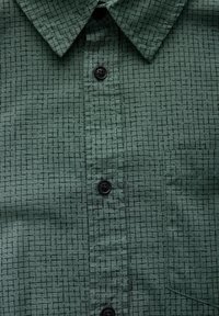 Zelená košile s knoflíky vyrobená z texturované látky s černým mřížkovaným vzorem, s špičatým límcem a jednou kapsou na hrudi.