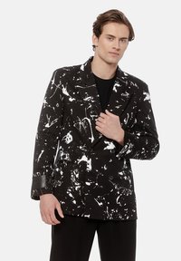 Młody mężczyzna w czarnym dwurzędowym żakiecie z białym wzorem w plamki farby, w połączeniu z czarnymi spodniami i czarną koszulą.