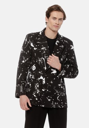 Młody mężczyzna w czarnym dwurzędowym żakiecie z białym wzorem w plamki farby, w połączeniu z czarnymi spodniami i czarną koszulą.