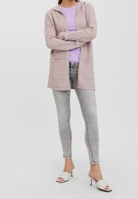 Světle růžový kardigan s kapucí a předními kapsami, doplněný lila topem, šedými skinny jeans a bílými sandály na podpatku.