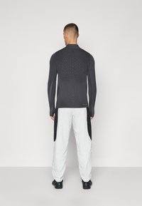 Dunkelgraues Langarmshirt mit strukturiertem Rippstrick, kombiniert mit weißen Hosen mit schwarzen Seitenpanelen. Das Model ist von der Kamera abgewandt.