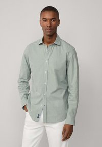 Camicia a maniche lunghe a quadri verdi e bianchi con colletto abbottonato, bottoni semplici e un dettaglio del logo sull'orlo, indossata con pantaloni bianchi.