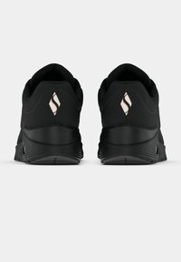 Schwarze Sportschuhe mit strukturierter Oberfläche, einem Logo an der Ferse und einer gerippten Sohle für besseren Halt. Rückansicht zeigt ein schlankes Design.