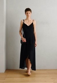 Abito nero senza maniche con scollo a cascata, orlo asimmetrico e una texture liscia, abbinato a una piccola borsa nera.