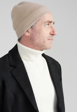 Beige gebreide beanie met een geribde boord, gecombineerd met een witte kabelgebreide coltrui en een zwarte jacket. Profielweergave.