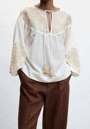 Hvid bluse med beige blomsterbroderi på ærmer og bryst, kvastbånd ved halsudskæringen, kombineret med løse brune bukser.