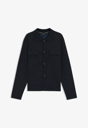 Zwarte blouse-jas met lange mouwen, knopen en een kraag, met twee borstflapzakken, plat gelegd op een witte achtergrond.