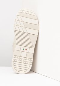 Suola di una scarpa crema con grip in gomma testurizzata, caratterizzata da motivi geometrici e branding. Dettagli sottili dei lacci visibili sul lato.