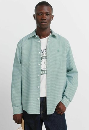 Jonge man met een groen geruit overhemd over een wit T-shirt met een afbeelding, donkere spijkerbroek, en een beige pet in zijn linkerhand.