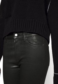 Jeans ajustados negros y brillantes con una textura suave y un cierre de botón visible. Combinados con un suéter negro de punto.