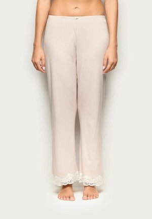 Pantaloni beige larghi con bordo in pizzo all'orlo indossati da una persona in piedi, visibile dalla vita ai piedi su uno sfondo semplice.