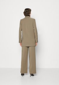 Sacou din material taupe, croit pe măsură, cu o siluetă lungă și pantaloni evazați. Tessut neted, design cu un singur nasture și detalii hardware minime.