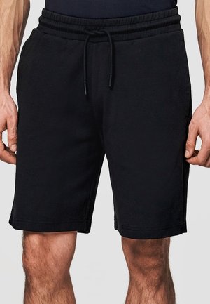 Czarne krótkie spodenki o średniej długości z regulowanym sznurkiem w talii, noszone przez stojącą osobę, na tle jednolitego tła, ukazujące dłonie i nogi.