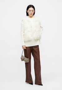 Colete de pelúcia branca sobre um suéter de malha creme, combinado com calças largas de imitação de couro castanho e uma pequena bolsa de ombro metálica.