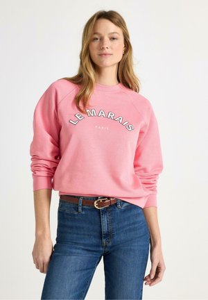 Nainen, joka pukeutunut vaaleanpunaiseen collegepaitaan, jossa on teksti "LE MARAIS PARIS", sinisiin farkkuihin ja ruskeaan vyöhön, seisoo yksivärisen valkoisen taustan edessä.