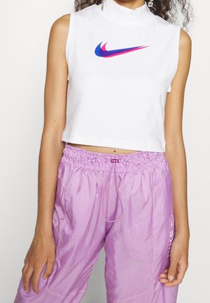 Femme portant un crop top blanc sans manches avec un logo Nike bleu et rose, ainsi qu'un pantalon Nike violet ample avec taille élastique et cordon de serrage.