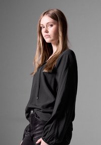 Black long-sleeve blouse with a drawstring neckline, made of soft fabric, featuring a relaxed fit and subtle balloon sleeves, paired with dark jeans.