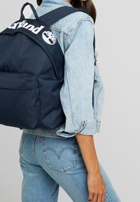Personne portant une veste en jean bleu clair et un jean, avec un sac à dos Timberland bleu marine portant le logo dans le dos.