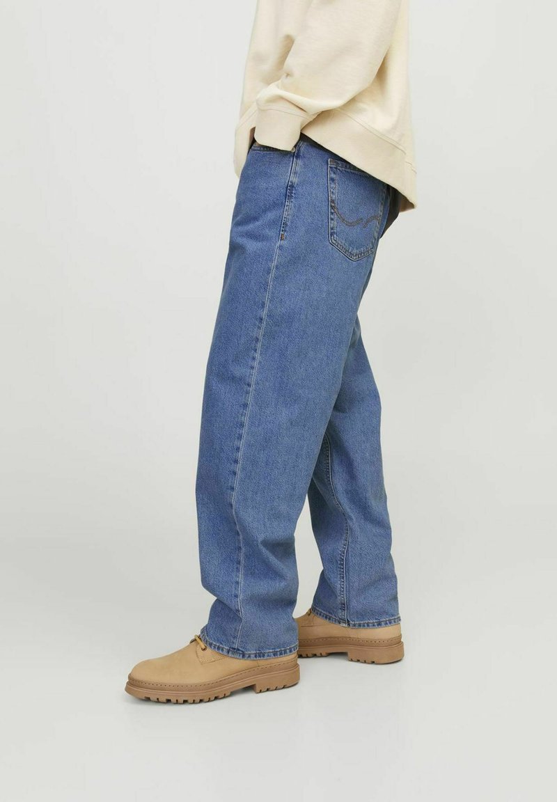 Osoba ubrana w luźne niebieskie dżinsy, beżowe buty sznurowane i kremowy sweter, stojąca na tle gładkiego, jasnego tła.